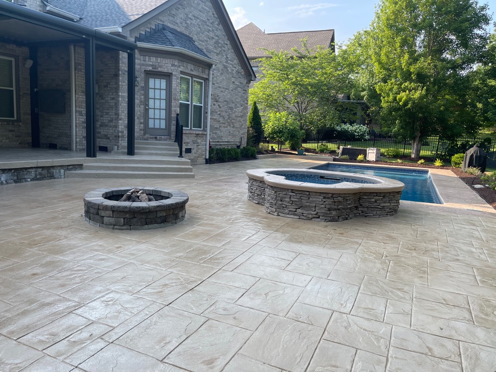 Stamped pool deck at a Brentwood home resurfaced to an even warm cream, running from the covered porch past a stone fire pit and raised spa